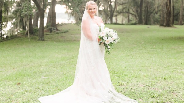 Bride in white gown holding bouquet, standing on grassy field with trees in background.