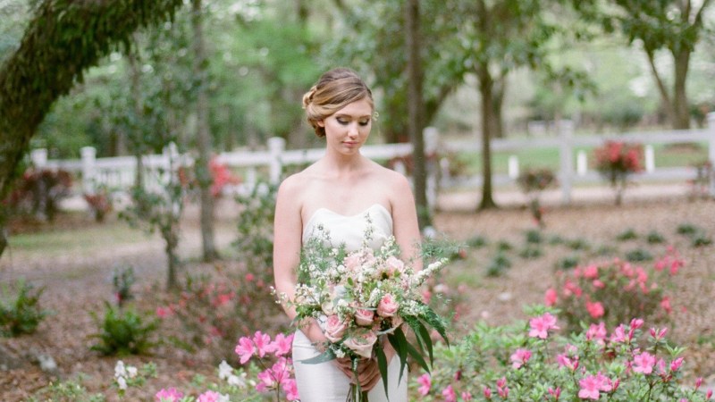 Bride with bouquet Bride in white dress holding bouquet stands in a garden with pink flowers and trees.