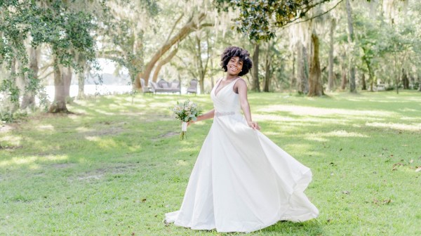 Bride in white dress holding bouquet, spinning on green grass among trees in sunlight.