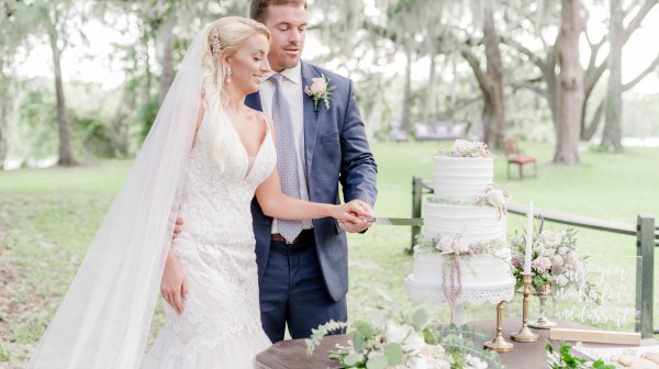 Bride and groom cutting a three-tiered wedding cake outdoors with floral decorations.