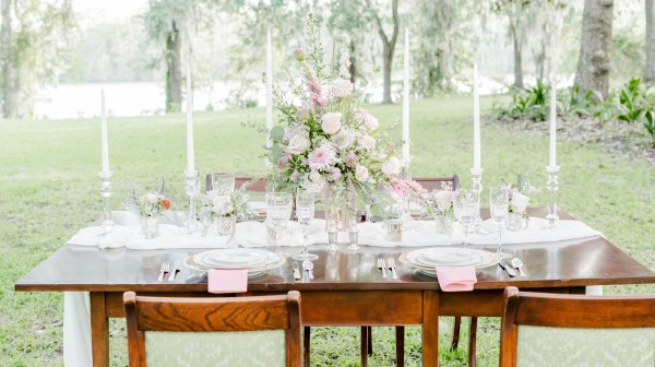 Outdoor table with flowers, candles, and plates on grass, surrounded by trees.