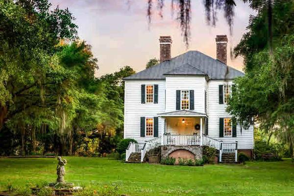 White colonial-style house with green shutters, surrounded by trees and a statue on the lawn.