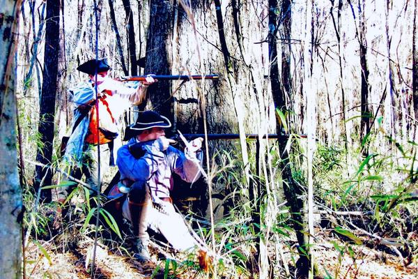 Two people in historical attire aiming rifles in a forest.