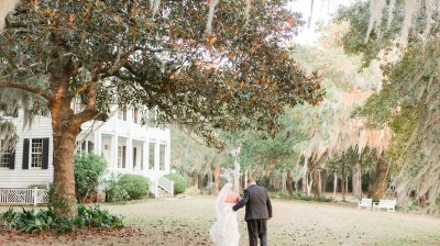 Bride and groom walking by a large tree near a white house on a grassy path.