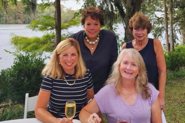 Four women gathered around a table with drinks by a lakeside.