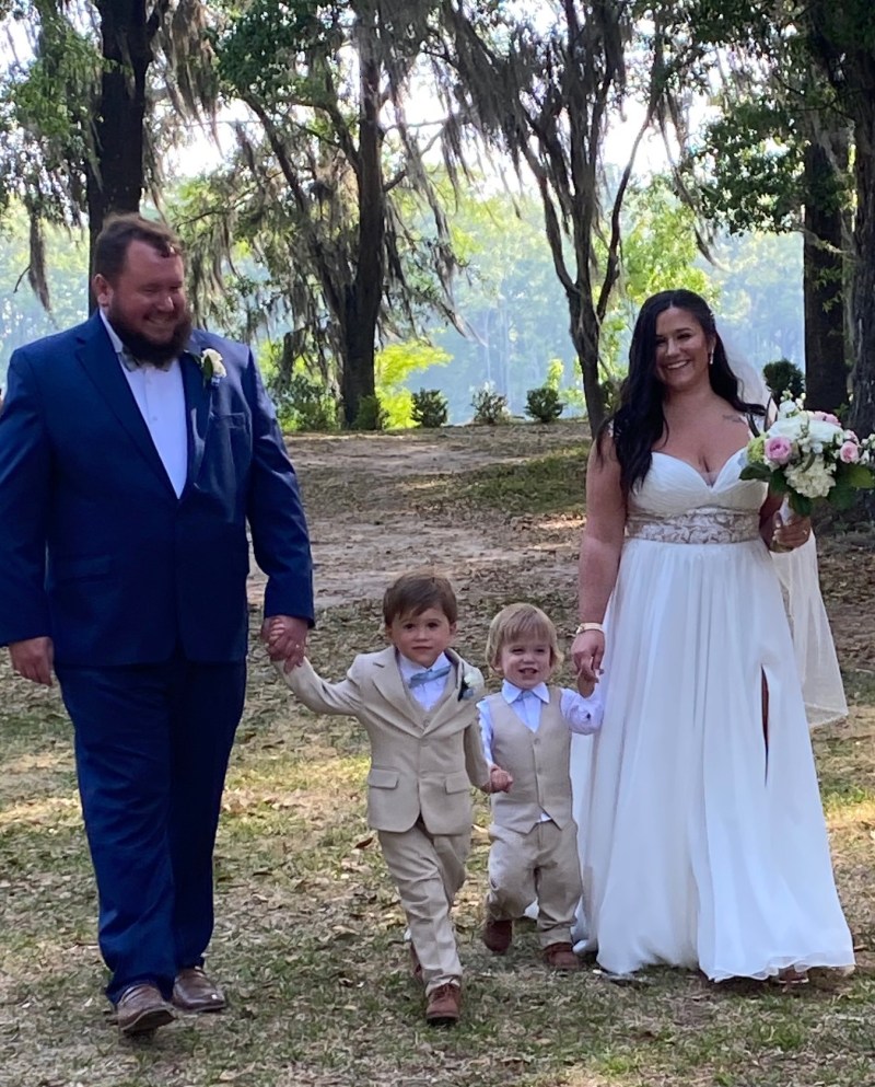 Couple in formal attire walking with two young boys in matching suits outdoors.