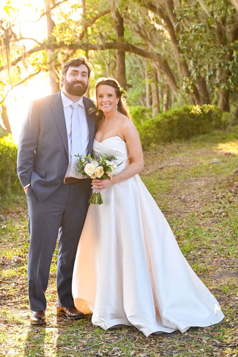 Bride and groom pose outdoors in formal attire on a sunny day.