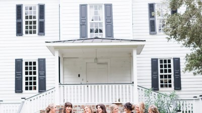 Bridal party in black dresses and bride in white, posing in front of a large white house.