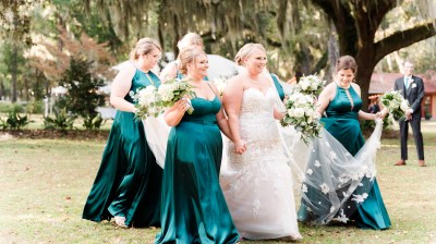 Bride in white dress with bridesmaids in teal gowns holding bouquets outdoors.