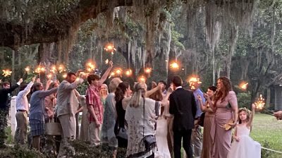 People walk through a forest path holding lit torches, surrounded by trees and greenery.