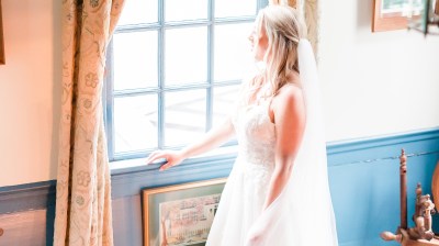 Bride in white dress stands near window, looking outside, with patterned curtains and decor around.