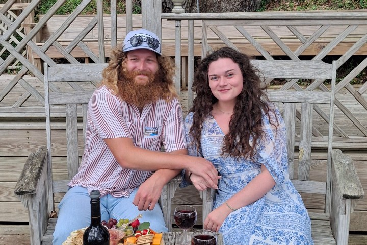 Couple seated on a bench with wine and cheese, outdoors on wooden deck.