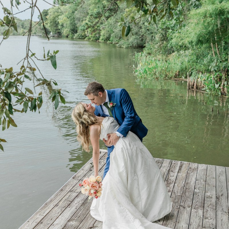 Couple in wedding attire kissing on a wooden deck by a lake.
