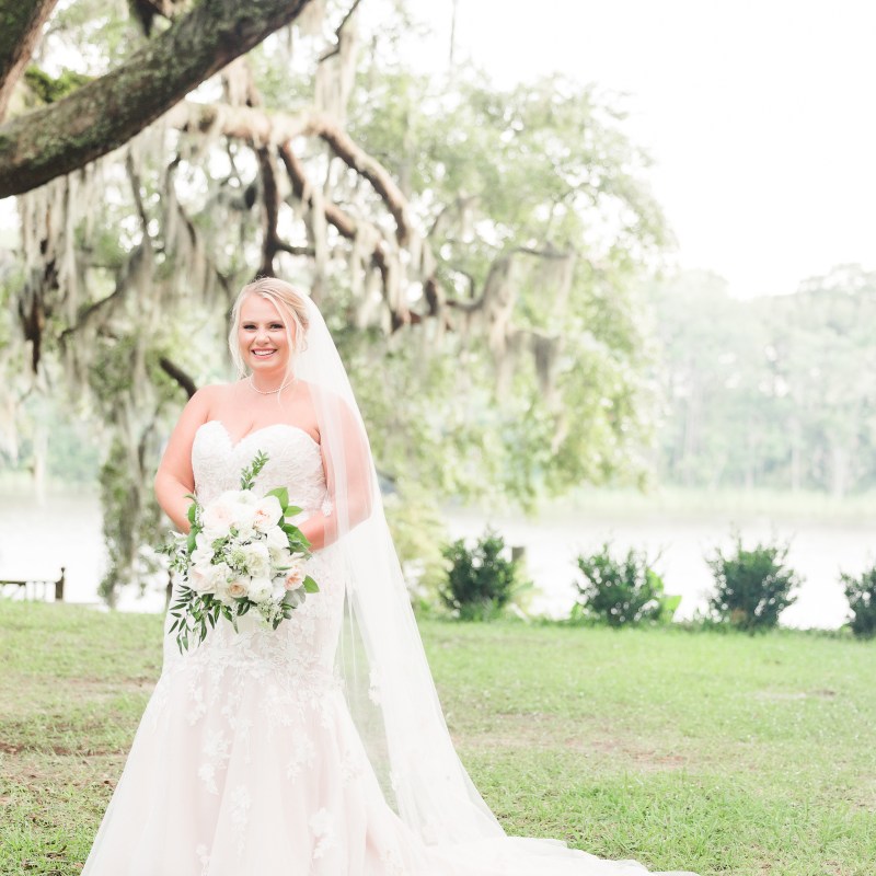 Bride in white gown holding bouquet under Spanish moss tree near a lake.