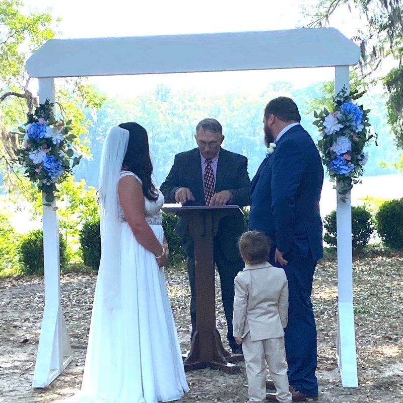 Wedding ceremony by a lake with couple, child, and officiant under floral arch.