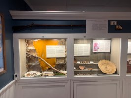 Museum display with artifacts, framed text, and a rifle mounted above.