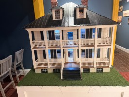 Model of a large white house with balconies, on a green base, in a display room.